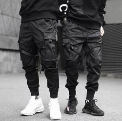 Men wearing black casual cargo pants with multiple pockets and edgy ribbon accents, styled in a modern hip hop fashion.