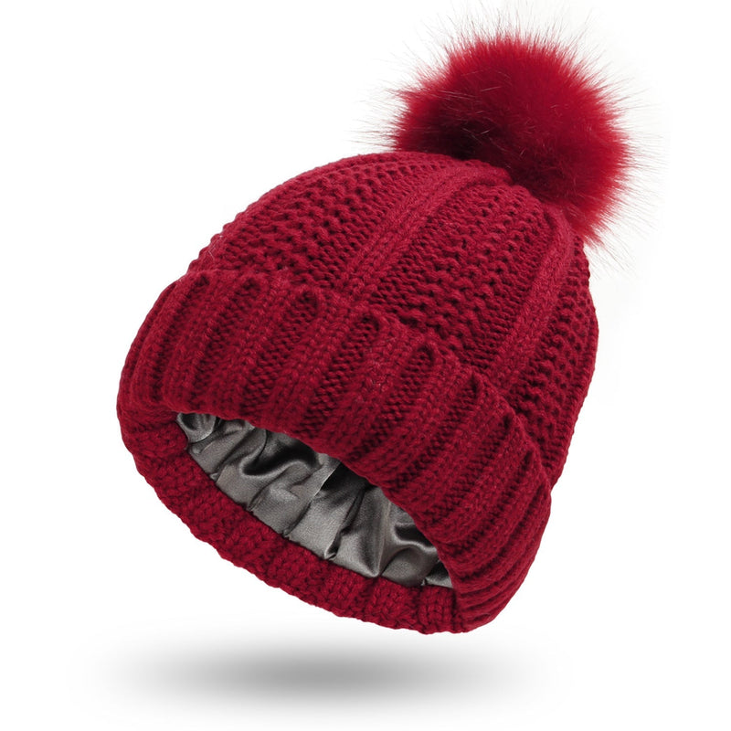 Satin-lined beanie with faux fur pom pom in wine red, knit design for warmth and hair protection.