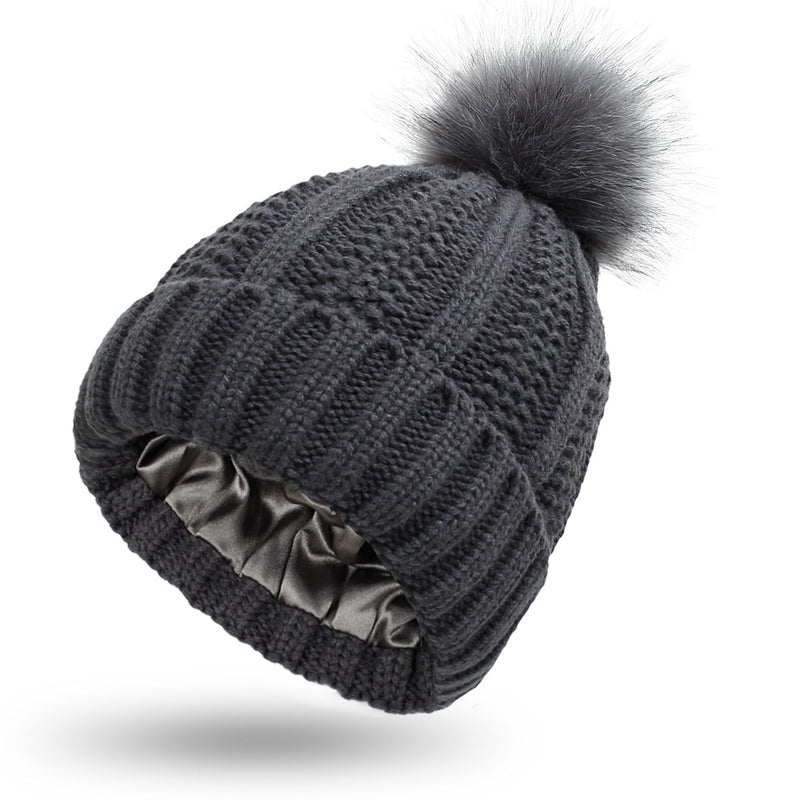 Satin-lined beanie with faux fur pom pom in dark gray, featuring knit design and silky inner layer for winter warmth.